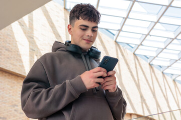 Young man using smartphone with headphones around neck in modern glass building, teenager texting...