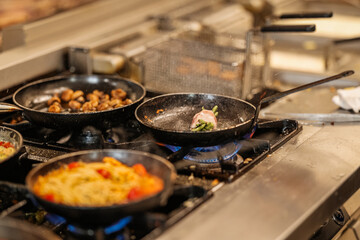 Close-up of sizzling pans on a restaurant gas stove. Focus on bacon-wrapped green beans, mushrooms,...