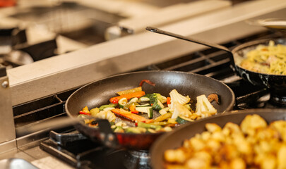 Colorful mixed vegetables being sautéed in a frying pan on a professional gas stove in a restaurant kitchen. Fresh ingredients and healthy cooking concept.