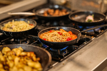 Spaghetti with tomato sauce and other dishes cooking in multiple pans on a gas stove. Close-up of hot food preparation in a busy professional kitchen environment.