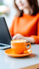 A vibrant orange coffee cup sits on a saucer atop a table, in focus, before a blurred woman using a laptop.  The latte art is visible on the frothy surface of the coffee