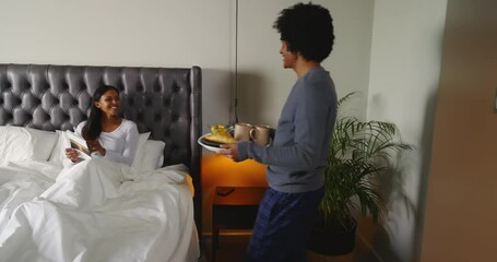Diverse couple sharing breakfast tray in bed with coffee mugs croissants after reading Sunsets book - Powered by Adobe