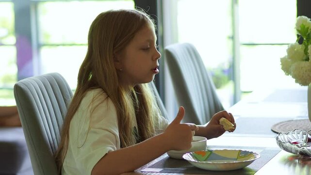 A girl is having breakfast at the kitchen table. A large window in the background. Healthy food for a child. The power supply mode. Children's diet. Healthy lifestyle. Rustic breakfast