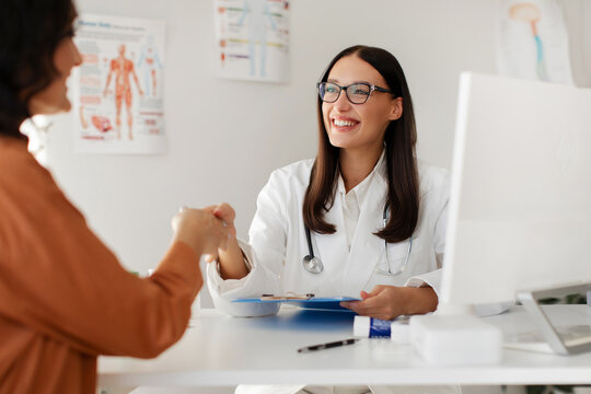 Medical services concept. Cheerful woman doctor and female patient handshaking during meeting in clinic, therapist welcoming lady
