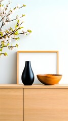 Minimalist home decor featuring a light wood sideboard, a black vase, a wooden bowl, a framed blank canvas, and a blossoming branch