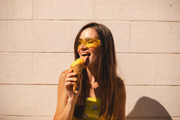 Close up happy young woman with delicious yellow ice cream in waffle cone outdoors. Girl wear yellow sunglasses in summer, spring or fall sunny day. Brunette female lick ice cream and laughing.