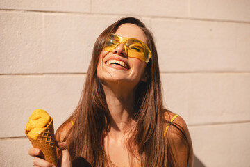 Close up happy young woman with delicious yellow ice cream in waffle cone outdoors. Girl wear yellow sunglasses and dress in summer, spring or fall sunny day. Brunette female laughing.