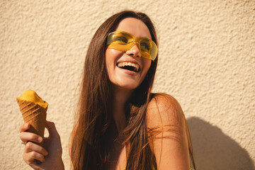 Brunette long hair woman eating an ice cream in waffle cone while standing in a street near wall. Happy female tourist wear yellow sunglasses, dress enjoying her summer vacation in a new city. 