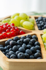 Close-up of assorted fresh berries and green grapes in wooden tray on table in natural light. Concept of healthy nutrition, summer fruits, organic food, clean eating, and vegan lifestyle.