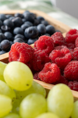 Close-up of fresh raspberries, blueberries and green grapes in wooden tray on kitchen table in natural light. Concept of healthy eating, summer fruits, organic food and plant-based nutrition.