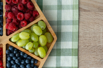 Fresh organic berries and grapes in wooden compartment box on rustic kitchen table with green checkered napkin. Healthy summer fruit assortment for breakfast or snack concept.