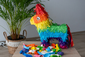 Colorful rainbow pinata with scattered balloons on wooden table next to green palm plant in modern home interior. Traditional Mexican party decoration for birthday celebration and festive events.
