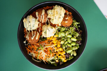 Falafel poke bowl with corn, edamame, greens, and creamy sauce, top view, isolated on green background.