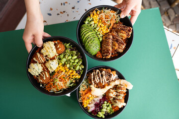 Three colorful poke bowls with falafel, avocado, grilled meat, and vegetables, top view, isolated on green background.