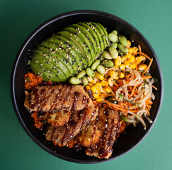 Poke bowl with sliced avocado, grilled meat, edamame, corn, and vegetables, top view, isolated on green background.