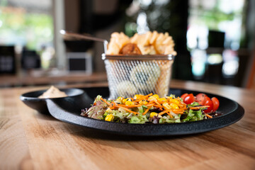 Close-up of waffle fries in metal basket with fresh salad and sauce on black plate, front view, isolated on wooden table.