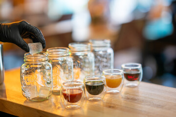 Gloved hand placing tea bag in mason jar with assorted drinks and jars, front view, isolated on wooden table background.