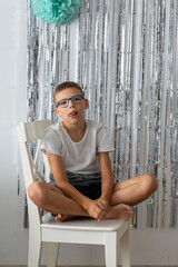 Cute boy with glasses sitting cross-legged on white chair against silver foil curtain backdrop with mint green paper pom-pom decoration. Portrait of child at birthday party or celebration setup.