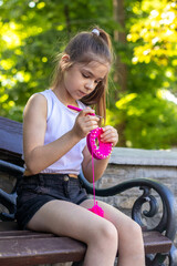 Young girl knitting with pink yarn on a park bench in summer