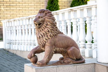 A stone lion statue with a mane, seated on a pedestal near white balusters and a brick wall