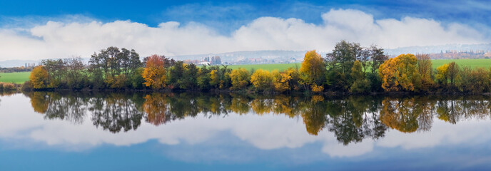 Autumn landscape with colorful trees reflecting in calm water, against a backdrop of fields and cloudy sky