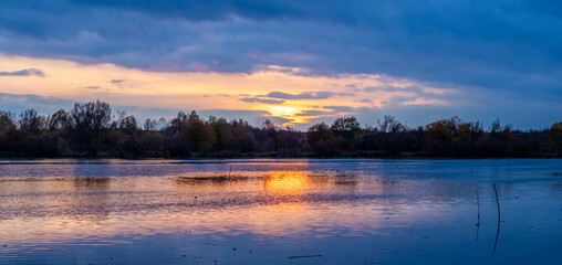 sunset over a calm river with fiery sky reflection in water, autumn landscape with tree silhouettes on the horizon