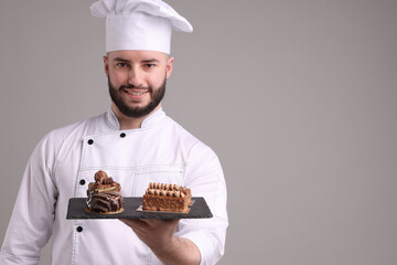 Happy confectioner in uniform holding delicious chocolate desserts on light grey background