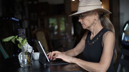 A mature woman sits at a cafe table, focused on her tablet as she enjoys a cup of coffee. The atmosphere is relaxed, perfect for digital nomads.