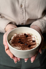 Holding a bowl filled with velvety dessert, layered with cream and cocoa, the warm kitchen light enhances the inviting atmosphere, perfect for a cozy gathering or sweet indulgence