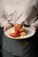 A smiling chef presents a beautifully plated pastry, drizzled with savory tomato sauce and sprinkled with nuts, radiating warmth and comfort. The inviting atmosphere enhances the delightful dish