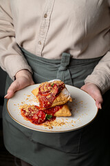 A skilled chef presents a beautifully arranged plate featuring stacked pastries drizzled with bright red sauce and sprinkled with crunchy crumbs. The warm atmosphere adds to the indulgent experience