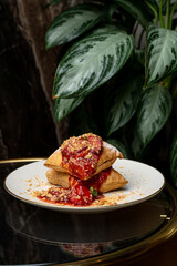 A stack of golden fried pastries is drizzled with a rich, red sauce and sprinkled with fresh herbs, resting on a chic plate against a lush green plant backdrop