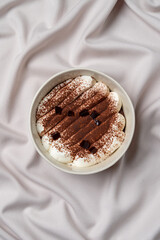 A decadent dessert features rich layers of creamy white frosting and cocoa powder, artfully arranged in a circular dish. Elegantly set against soft fabric, it invites enjoyment