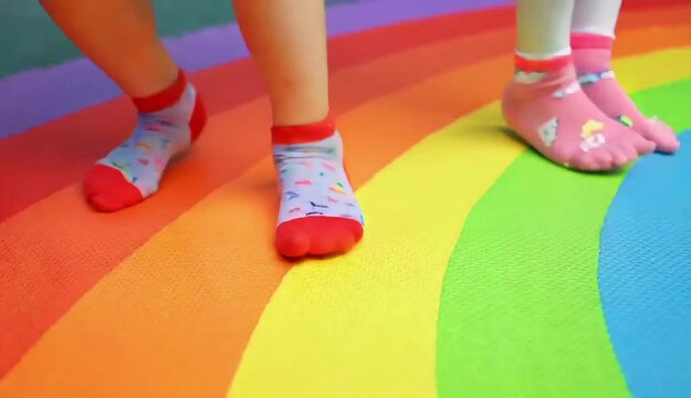 Close-up shot of two children's feet, tiny socks, rhythmically tapping on a vibrant rainbow-colored floor mat. Their small legs dangle playfully as they tap in time, generating a happy, childlike ener