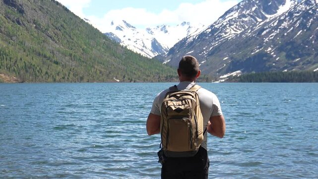 Man traveler in the mountains. Hiker with a backpack reached a mountain lake, the man poses against the background of the lake and snowy mountain peaks in the distance.