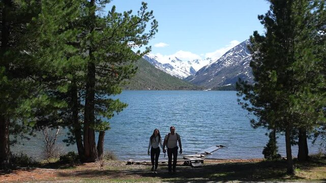 Mountain travel. A couple with backpacks reached a mountain lake, snowy peaks in the distance. Mountain travel and hiking. A couple posing against the backdrop of a lake and mountains