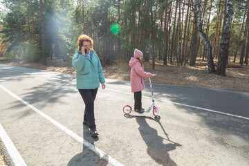 Grandmother talking on the phone while walking with her granddaughter who is riding a scooter in...