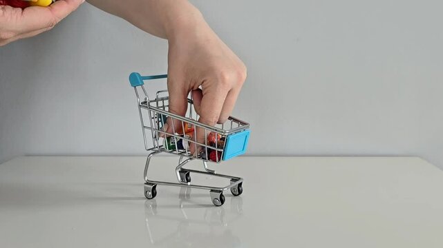 A hand takes out toy groceries from a mini shopping cart on a white table with a gray background &mdash; symbolizing family food shopping, rising prices, and everyday expenses.