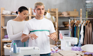 Careful young Asian tailoress measuring male client using tape in sewing workshop