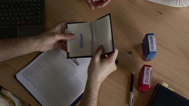 Top down shot of hands of unknown male officer stamping passport of female applicant approved and giving it back to her during workday at wooden desk in visa center