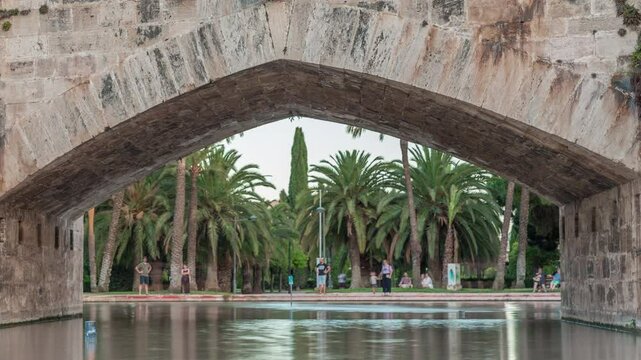 Arch of Pont de la Mar bridge timelapse in Valencia, Spain, featuring its ornate architecture reflected in a tranquil pond. Surrounded by green parkland and palm trees under a clear blue sky.