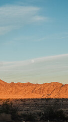 Naklejka premium Desert Mountains Illuminated by Sunset with Moon Rising in Soft Blue Sky — Dramatic Landscape of Arid Terrain, Warm Evening Light, and Celestial Scene for Nature, Travel, and Minimalist Photography