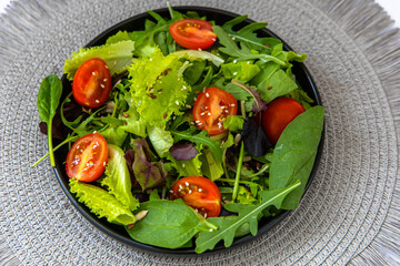 Fresh green salad with cherry tomatoes and seeds served in a black bowl on a textured placemat