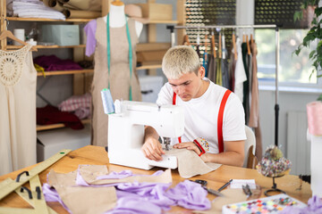 Young man tailor sews clothes on a sewing machine in the workshop. Fashion designer is working on...