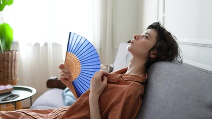 Exhausted woman lying on sofa soaked in sweat using fan to relieve extreme heat. Heatstroke risk, indoor climate discomfort, physical fatigue, urban summer survival, dehydration, no conditioning - Powered by Adobe