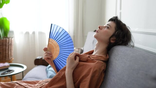 Exhausted woman lying on sofa soaked in sweat using fan to relieve extreme heat. Heatstroke risk, indoor climate discomfort, physical fatigue, urban summer survival, dehydration, no conditioning