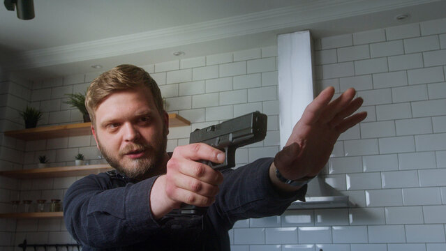 Bearded man points handgun with one hand while extending the other in a stop gesture, suggesting a tense or defensive situation.