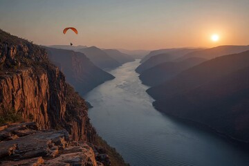 Fototapeta premium A paraglider soars above a river canyon at sunset, with dramatic rock cliffs, layered mountains and a golden sky creating a breathtaking and adventurous scene.
