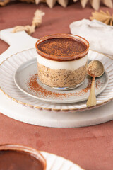 Tiramisu chia pudding in a glass cup on a white plate with a spoon, garnished with cocoa powder, set on a brown surface with a sieve and cocoa powder in the background