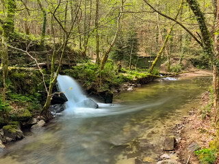 A scenic view of Bugio River's natural waterfall cascading amidst lush greenery and trees, with a small stone structure nearby in Jugueiros, Felgueiras, Portugal